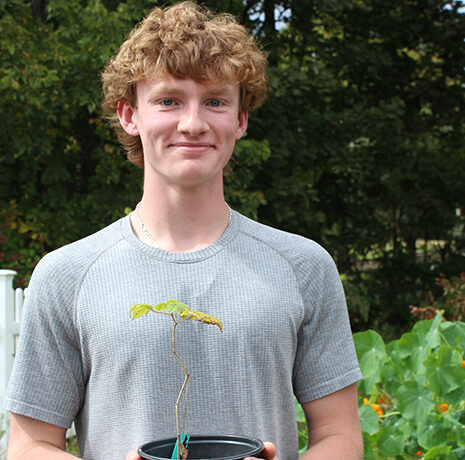 A young person with curly hair stands outside in a garden, holding a small potted plant. A white fence and green plants are visible in the background.