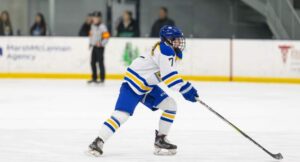 A hockey player in blue and white uniform skates on the ice with a stick, preparing to play, while officials and spectators watch in the background.