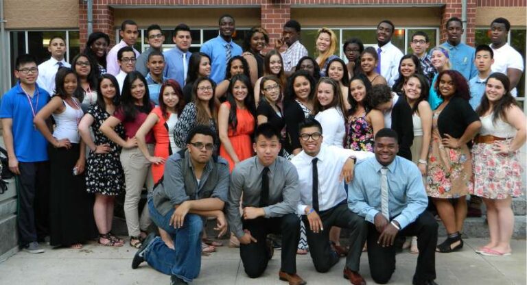 A large group of young adults pose together outside a building, some standing and some crouching, dressed in business casual and semi-formal attire.