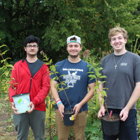 Three young men stand outdoors, each holding a potted plant, with greenery and trees in the background.