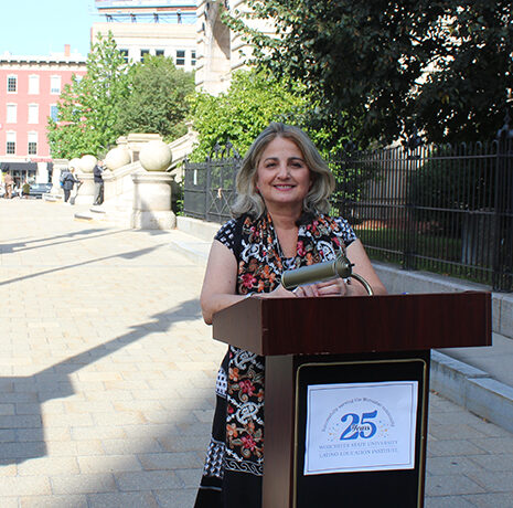 A woman stands at a podium with a microphone outdoors, smiling. The podium displays a sign celebrating 25 years of the Columbia Center for Oral History Research.