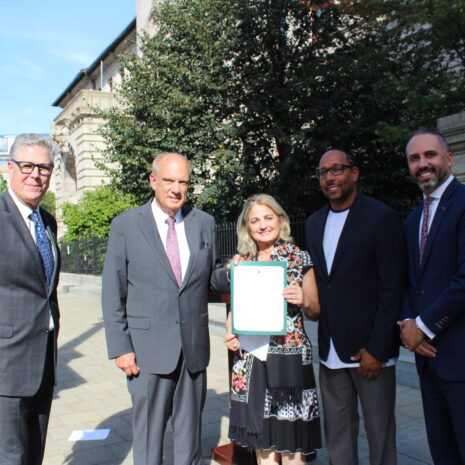 Five people stand outdoors in business attire, with one woman in the center holding a certificate; buildings and greenery are visible in the background.