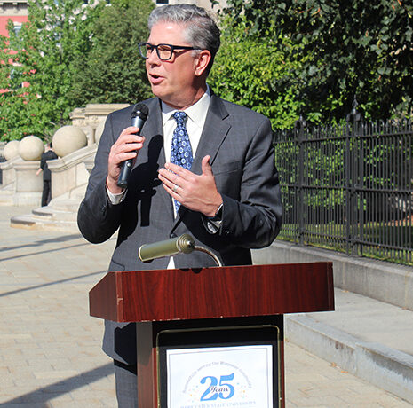 A man in a suit speaks into a microphone at an outdoor podium during a daytime event.