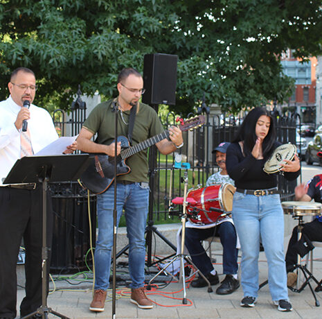 Five people perform music outdoors; one sings, one plays guitar, one holds a tambourine, and two play drums. They are standing and seated on a paved area near a park.