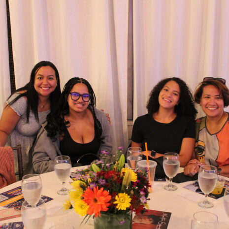 Four women sit at a round table set with water glasses and floral centerpiece, smiling at the camera in a well-lit room with white curtains.