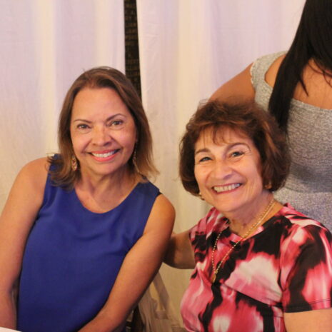 Two women are seated and smiling at the camera, while another person with long hair faces away in the background. The setting appears to be indoors with cream-colored curtains.