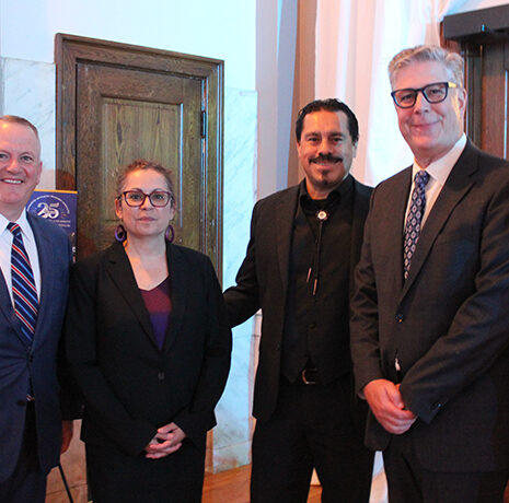 Five adults, three men in suits and two women in business attire, stand together indoors, posing for a group photo near wooden doors.