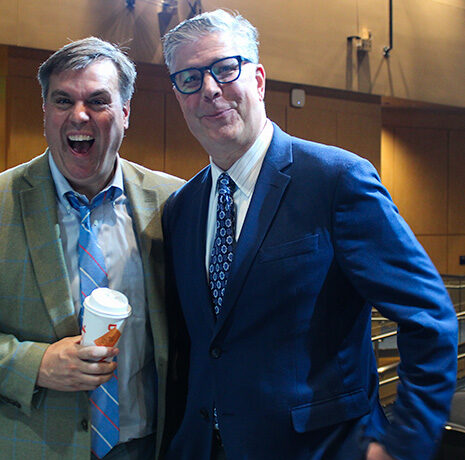 Two men in suits stand together in an auditorium; one is smiling widely while holding a coffee cup, the other is wearing glasses and smiling slightly.