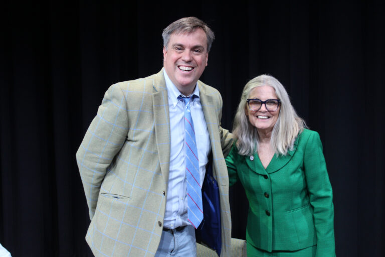 Two people, one man in a checked blazer and tie and one woman in a green suit, stand and smile in front of a black background.