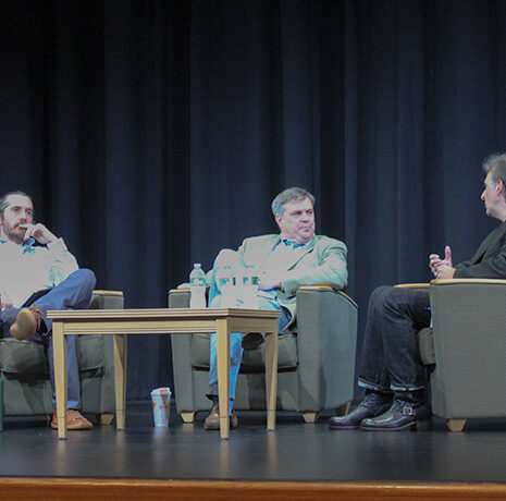 Three men sit and converse on stage in armchairs, with a table holding drinks between them, in front of a black curtain.
