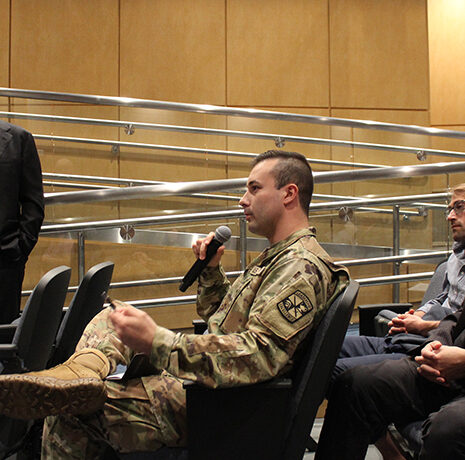 A man in military uniform speaks into a microphone while seated in an auditorium with three other men, one standing and two seated.