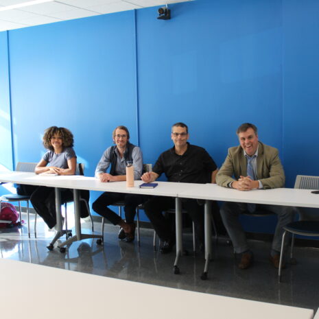 Five people sit at tables in a brightly lit room with blue walls, facing the camera and smiling. Laptops, notebooks, and bags are visible on the tables.