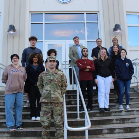 A group of twelve people, including one in military uniform, stand on the steps outside a building with large windows and double doors.