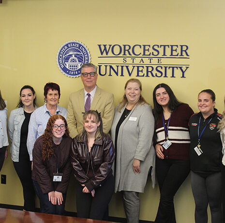 A group of ten people, some seated and some standing, pose in front of a Worcester State University logo on a yellow wall.