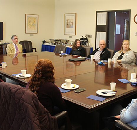 A group of people sit around a large conference table having a meeting; coffee cups, plates, and napkins are on the table.