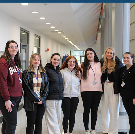 Seven women stand in a hallway, facing the camera and smiling. They are wearing casual clothing and ID badges. The hall is modern with bright lighting and several doors visible.