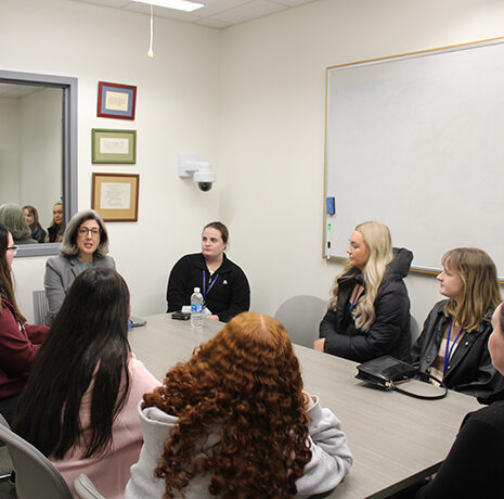 Seven women sit around a rectangular table in a small conference room, having a discussion. A whiteboard and framed certificates are visible on the walls.
