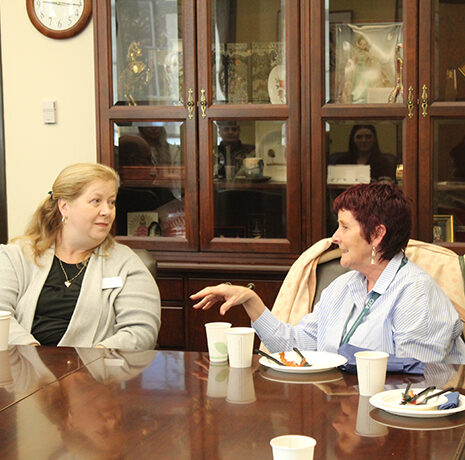 Two women sit at a table talking, with plates and cups in front of them. A glass cabinet with various items is in the background.