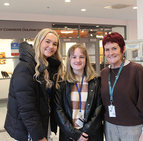 Three women stand together indoors, smiling at the camera. They are wearing work lanyards and jackets, with a gallery entrance visible in the background.
