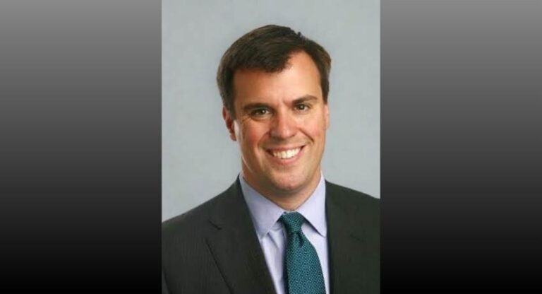 A man with short dark hair wearing a dark suit, light shirt, and blue patterned tie, smiling at the camera against a plain background.