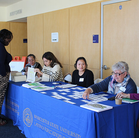 Five women sit at a table with a blue “Worcester State University Latino Education” tablecloth, checking in a visitor in a hallway with wooden walls and doors.