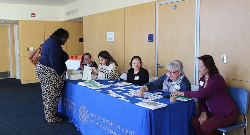 Five women sit at a table with a blue “Worcester State University Latino Education” tablecloth, checking in a visitor in a hallway with wooden walls and doors.
