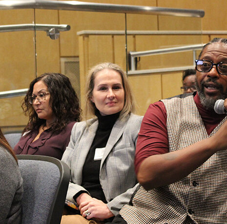 A man holding a microphone speaks while sitting among a group of people in an auditorium or conference room.