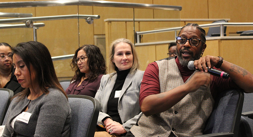 A man holding a microphone speaks while sitting among a group of people in an auditorium or conference room.