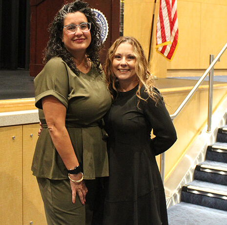 Two women stand and smile for a photo in an auditorium, with an American flag and carpeted stairs visible in the background.