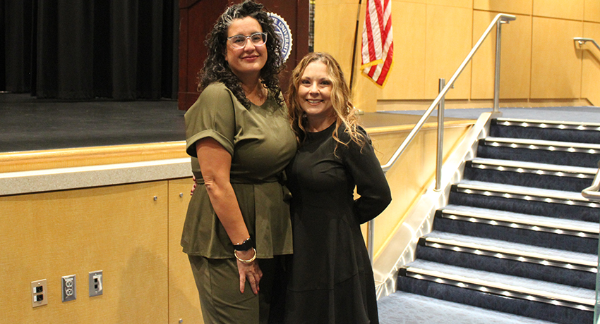 Two women stand and smile for a photo in an auditorium, with an American flag and carpeted stairs visible in the background.