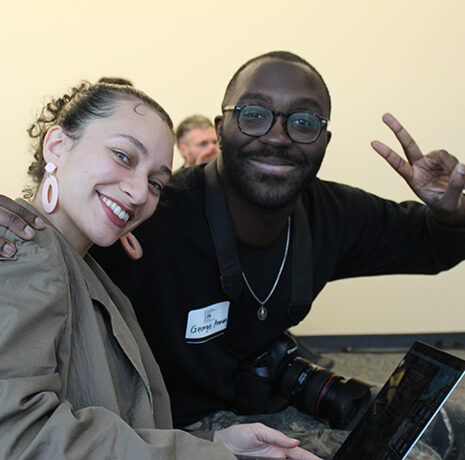 Two people sit together indoors, smiling at the camera; one person holds up a peace sign and a camera rests on their lap.