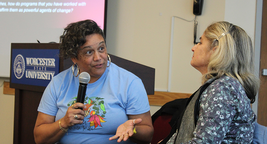 Two women are engaged in conversation in front of a Worcester State University podium; one gestures while speaking.