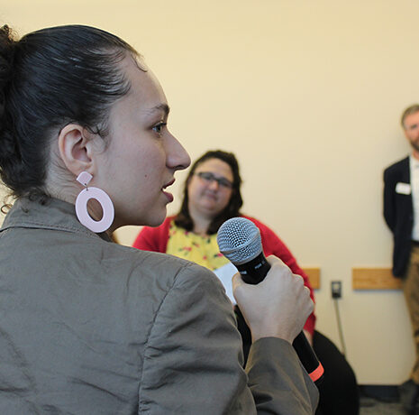 A woman holding a microphone speaks at an indoor event, while two people listen in the background.