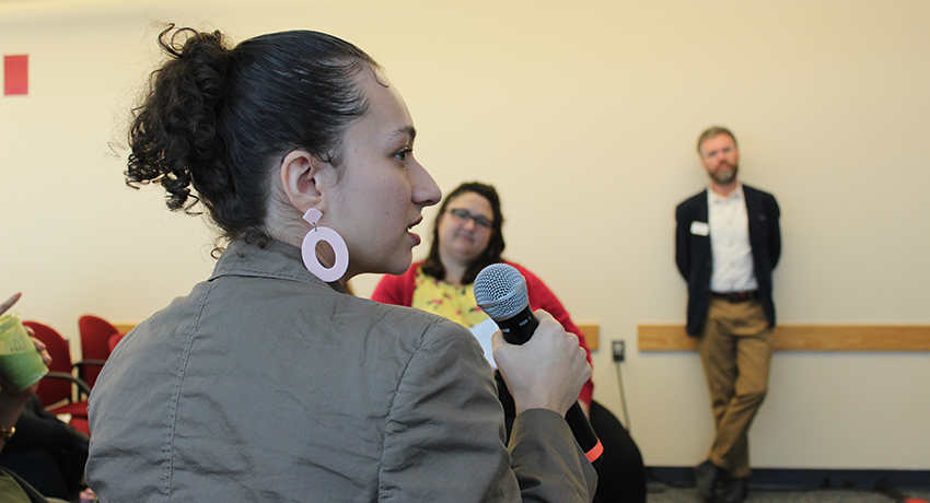 A woman holding a microphone speaks at an indoor event, while two people listen in the background.