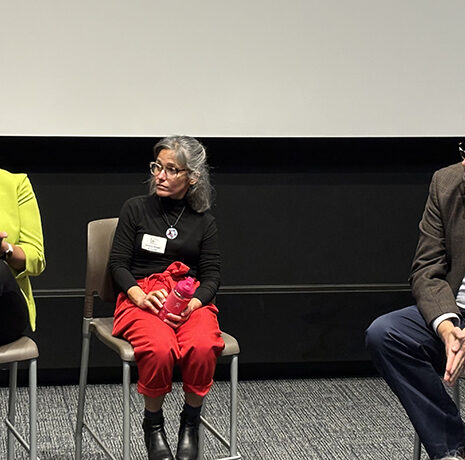 Three people sit on chairs in front of a white screen, engaged in conversation during what appears to be a panel discussion or event.