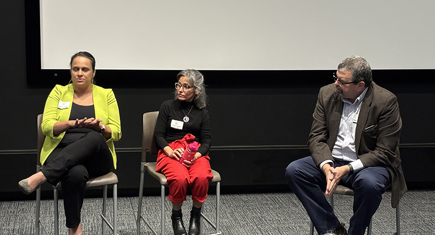 Three people sit on chairs in front of a white screen, engaged in conversation during what appears to be a panel discussion or event.