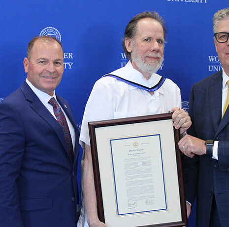 Three men pose in front of a Worcester State University backdrop; the man in the center holds a framed certificate or award.