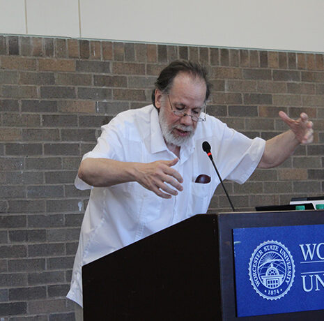 An older man gestures while speaking at a podium with a Worcester State University sign in front, in a room with brick walls.