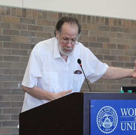 An older man with glasses speaks at a podium labeled 