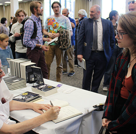 A man sits at a table signing a book for a woman while a line of people waits behind her at an indoor event.