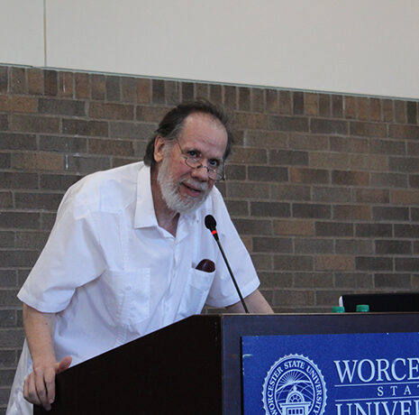 An older man with glasses and a white shirt speaks at a podium labeled 