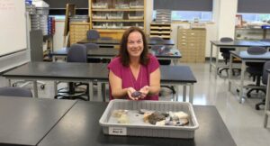 A woman sits at a classroom table, smiling and holding rocks, with a tray of geological samples in front of her. Shelves and cabinets are visible in the background.