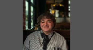 A person with short curly hair, wearing a striped shirt and bolo tie, smiles while sitting at a restaurant booth near a window.