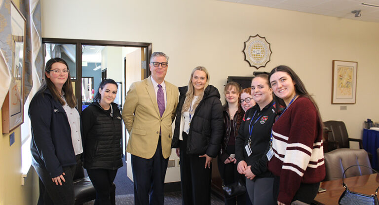 A group of seven people, including one man in a tan blazer, stand together indoors, smiling at the camera in an office-like setting.