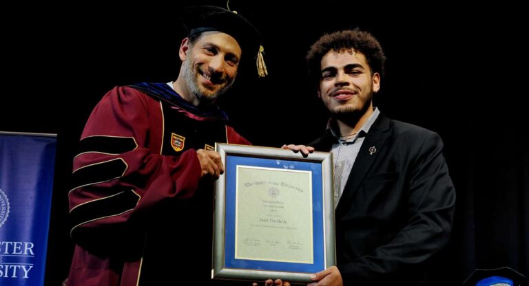 Two men stand side by side, smiling and holding a framed certificate during an award ceremony. One is wearing academic regalia, and the other is in a suit.
