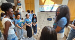 A group of students stand around a large research poster while one student presents findings in a brightly lit indoor space.