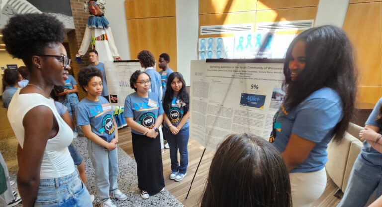A group of students stand around a large research poster while one student presents findings in a brightly lit indoor space.