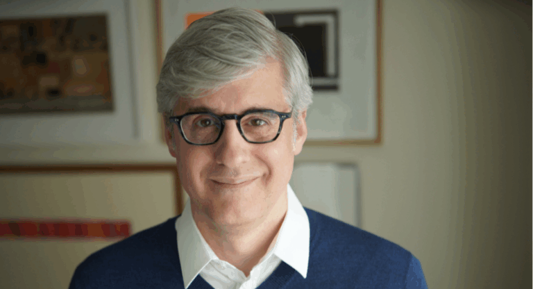 A man with gray hair and glasses, wearing a white collared shirt and blue sweater, stands indoors in front of framed artwork.
