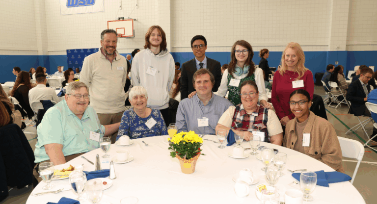A group of nine people pose for a photo around a round table with plates, cups, and a yellow potted flower at a formal event in a gymnasium.