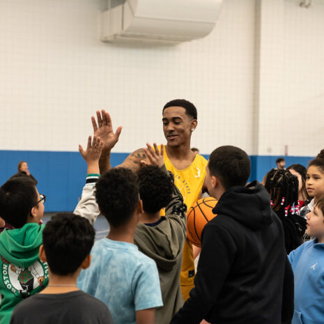 A basketball player in a yellow jersey smiles and high-fives a group of children in a gymnasium.